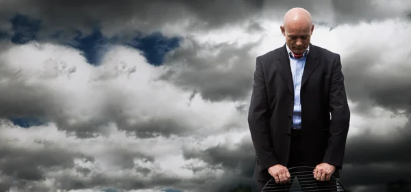 Mann im Anzug mit demütig gesenktem Kopf vor wolkenschwerem Himmel stützt sich auf einen Grillrost