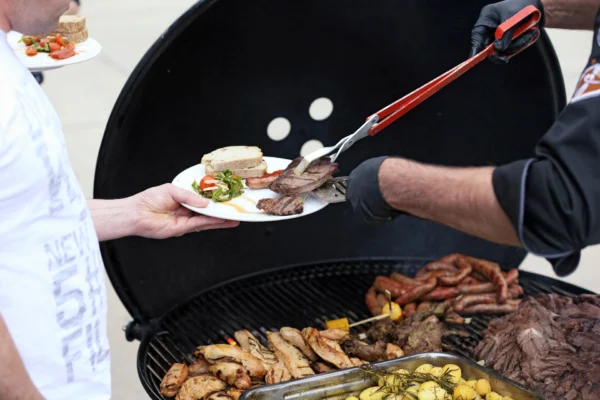 Weber Ranch Kettle Grill mit geöffnetem Deckel auf dem Rost liegen Grillwürste Fleisch und Gemüse
